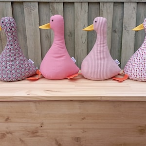 Könnte beinhalten: Vier Plüsch-Enten in einer Reihe. Die Enten sind aus verschiedenen Stoffen gefertigt und haben unterschiedliche Muster. Die Enten sind rosa, rosa gestreift und zwei mit einem rosa-weißen geometrischen Muster. Die Enten haben orangefarbene Schnäbel und Füße.