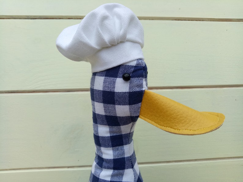 May include: A blue and white checkered fabric duck with a white chef's hat and a yellow leather beak.
