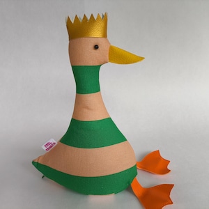 Peut inclure: Une peluche en forme de canard avec une couronne dorée, un bec jaune et des pieds orange. Le corps du canard est beige avec des rayures horizontales vertes. Une petite étiquette est attachée au corps du canard.