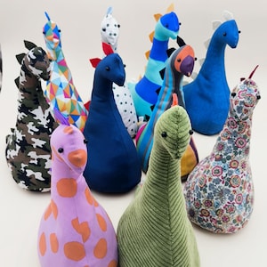 Könnte beinhalten: Eine Sammlung farbenfroher, gefüllter Dinosaurier-Türstopper. Die Türstopper weisen verschiedene Muster auf, darunter Camouflage, Polka Dots und Blumenmuster. Jeder Dinosaurier hat ein einzigartiges Farbschema mit Details wie Stacheln und Hörnern.