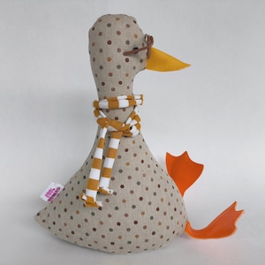Puede incluir: Un juguete de pato de peluche con un cuerpo beige con lunares, patas naranjas y un pico amarillo. Lleva una bufanda a rayas y gafas pequeñas. El pato está sentado, con las patas extendidas.