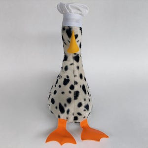 Peut inclure: Une peluche en forme d'oie dalmatien, portant un chapeau de chef blanc et un bec jaune. L'oie a des taches noires sur un corps blanc et des pieds orange. Ce jouet en peluche est conçu pour la décoration.
