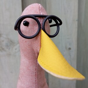May include: A pink fabric bird with a yellow beak and black round glasses. The bird is wearing a pair of black round glasses.