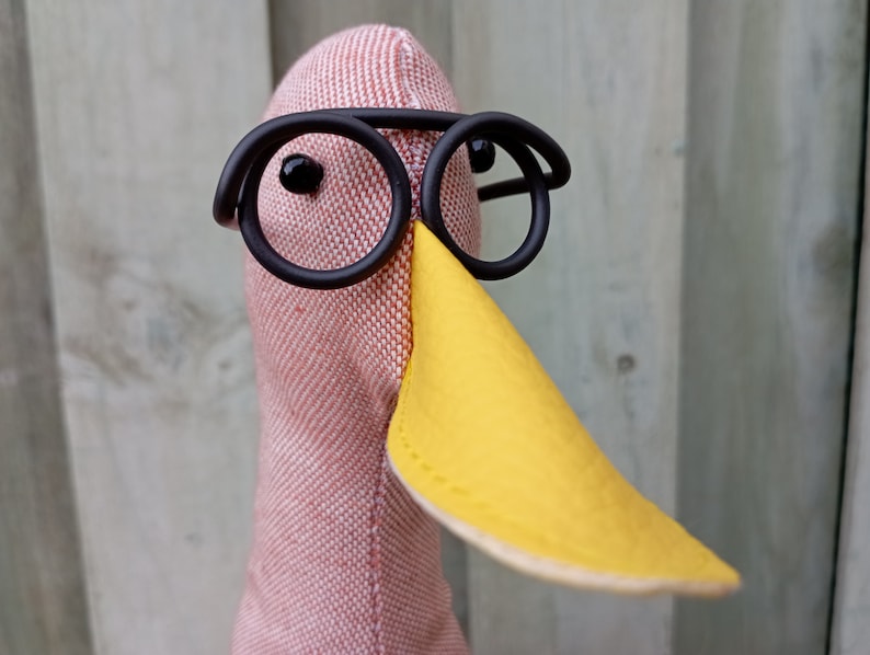 May include: A pink fabric bird with a yellow beak and black round glasses. The bird is wearing a pair of black round glasses.