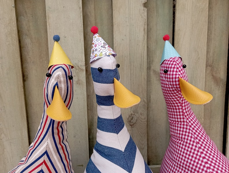 May include: Three plush toy ducks wearing party hats. The ducks are striped in red and white, blue and white, and red and white gingham.