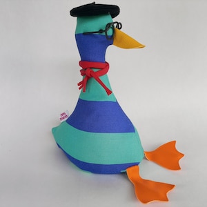 Peut inclure: Un canard en peluche fantaisiste avec un béret noir, des lunettes et une écharpe rouge. Le canard présente des rayures bleues et turquoise, un bec orange et des pieds orange. Une petite étiquette est visible sur le côté.