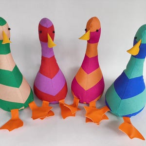 Puede incluir: Cuatro juguetes de pato de tela coloridos. Cada pato tiene un cuello largo y un pico amarillo. Los patos son de varios colores, incluyendo verde, rosa, naranja y azul, con patas palmeadas naranjas.