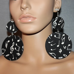 May include: Black and white musical note earrings. These large, circular earrings feature a black background with a repeating pattern of white musical notes, treble clefs, and other musical symbols. The earrings are suspended from small hooks.