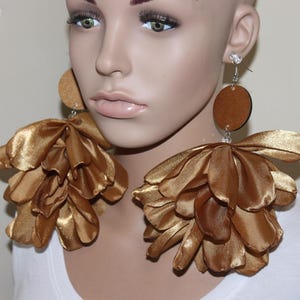 May include: Gold-colored statement earrings. Each earring features a large, layered flower design made of a shiny fabric. The earrings also have a small, round, brown accent piece at the top, and are suspended from a silver-colored hook.