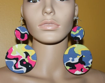 May include: A pair of large, round earrings with a colorful abstract pattern in shades of blue, pink, yellow, black, and white. The earrings are made of fabric and have a silver hook closure.