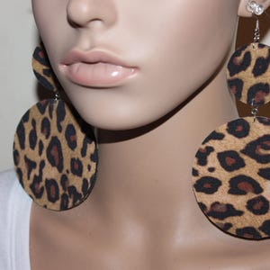 May include: Large, round leopard print earrings. The earrings feature a tan background with dark brown spots. The earrings are made of a lightweight material and hang from silver-colored hooks. The earrings are a bold fashion statement.