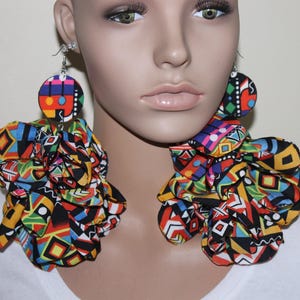 May include: Oversized drop earrings featuring circular fabric-covered studs attached to large, voluminous fabric ruffles. The fabric displays a vibrant, multi-colored geometric pattern in shades of red, yellow, blue, green, black, and white.