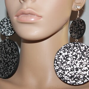 May include: Large, dangling earrings with a black and silver speckled pattern. The earrings are composed of three circular pieces of varying sizes, suspended from silver-colored hooks. The design features a bold, abstract aesthetic.