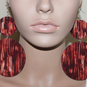 May include: Large, dangling earrings with a circular design. The earrings feature a two-tiered design with a smaller circle above a larger circle. The surface has a reddish-brown pattern with vertical streaks, and the earrings are suspended from silver-colored hooks.