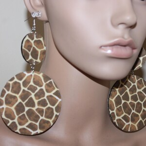 May include: Large, round earrings with a giraffe print pattern in shades of brown and tan. The earrings are suspended from silver-colored hooks and feature a smaller, matching disc above the larger circle.