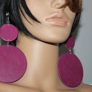 May include: A pair of large, magenta-colored wooden earrings. Each earring features two connected circular pieces, with the larger circle at the bottom. The earrings hang from silver-colored hooks. The earrings are a bold fashion statement.
