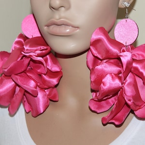 May include: Hot pink statement earrings. Each earring features a large, ruffled flower design made of satin fabric. The flowers are attached to a small, circular stud. The earrings are a bold and eye-catching accessory.
