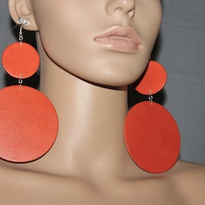May include: A pair of large, orange, round, dangle earrings. The earrings are made of leather and have a silver chain connecting the two circles.