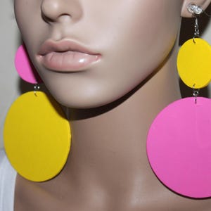 May include: Bright, oversized earrings with a retro vibe. The earrings feature a combination of pink and yellow circular shapes. The design includes a small pink circle, a yellow circle, and a large pink circle. The earrings are suspended from silver-colored hooks.