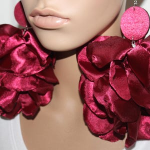 May include: Burgundy-colored statement earrings. Each earring features a large, layered flower design made of a silky fabric. The earrings have a circular stud at the top and hang down, creating a bold and dramatic look.