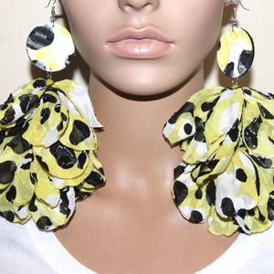May include: A pair of large statement earrings featuring circular tops with a yellow, black, and white animal print pattern. Attached to each top is a voluminous, layered cluster of translucent fabric petals in the same yellow, black, and white color scheme.