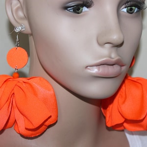 May include: Bright orange statement earrings. Each earring features a circular top, a small bead, and a large, layered flower design. The earrings are made of fabric and hang from silver-colored hooks.