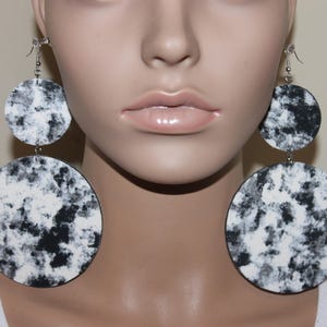 May include: Large, dangling earrings with a two-tiered circular design. The earrings feature a black and white marbled pattern. The top circle is smaller than the bottom circle. The earrings are suspended from silver-colored hooks.