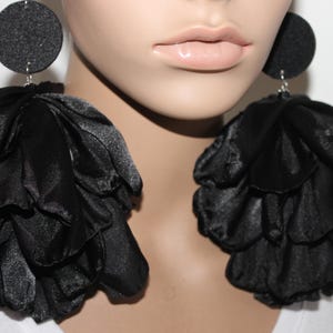 May include: A pair of statement earrings featuring a black circular stud top with a glittery texture. Attached to each stud is a large, voluminous cluster of layered black fabric petals that create a full, ruffled, and dimensional floral appearance.