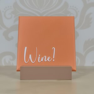 Puede incluir: Un letrero cuadrado naranja con la palabra "Wine!" en escritura blanca se muestra en un soporte marrón claro. El fondo presenta un papel pintado estampado.