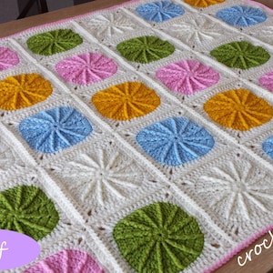 Pode incluir: Um cobertor de bebé em crochê com fundo branco e motivos circulares coloridos em rosa, amarelo, azul e verde. O cobertor tem uma borda rosa e o texto "crochets4U" é visível no canto inferior direito.