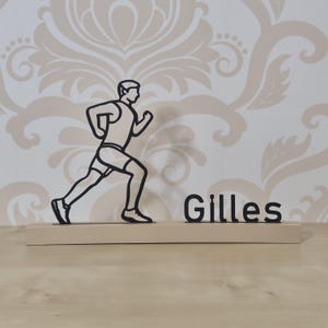 Op de afbeelding: Houten silhouet van een hardloper met de naam "Gilles" in zwart. De hardloper wordt afgebeeld in een rennende pose, gekleed in sportkleding. De basis is lichtbeige van kleur en de achtergrond heeft een bloemenpatroon.