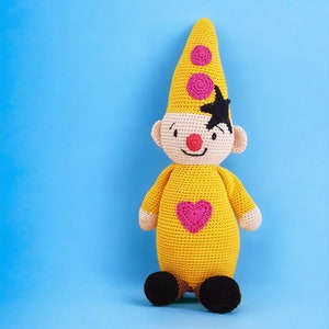 Op de afbeelding: Een gehaakt geel speelgoed met een roze hart op de borst, een zwart-wit gezicht en een roze-witte punthoed.