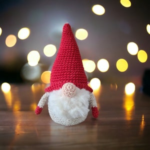 May include: A crocheted gnome with a red hat and white beard. The gnome is sitting on a wooden surface with a blurred background of lights.