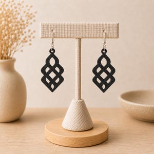 Op de afbeelding: Een paar zwarte oorbellen met een ingewikkeld, symmetrisch rasterpatroon. De oorbellen hangen aan zilverkleurige metalen haakjes en worden gepresenteerd op een beige, T-vormige stoffen sieradenstandaard met een ronde houten voet.