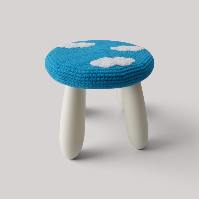K&ouml;nnte beinhalten: Ein kleiner Hocker mit einer runden Sitzfl&auml;che, die mit blauem H&auml;kelstoff bezogen ist und wei&szlig;e, geh&auml;kelte Wolkenmotive aufweist. Der Hocker hat drei glatte, abgerundete, cremewei&szlig;e Beine, die sich nach unten hin verj&uuml;ngen und f&uuml;r Stabilit&auml;t sorgen.
