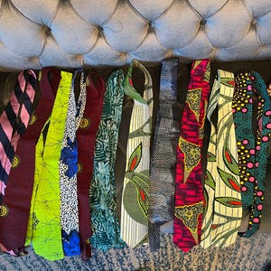 May include: A collection of colorful neckties with various patterns, including stripes, floral, and geometric designs. The ties are made of different fabrics and feature a range of colors, such as blue, green, red, and yellow. The ties are arranged on a gray upholstered surface.