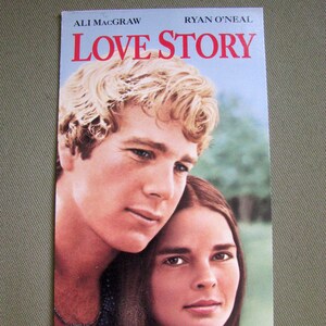 以下が含まれることがあります： アリ・マッグローとライアン・オニール主演の映画「愛と別れ」のヴィンテージ映画ポスター。ポスターは、アリ・マッグローがライアン・オニールの肩に寄りかかっている、2人の俳優のクローズアップをフィーチャーしている。