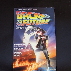 Puede incluir: Una carátula de cinta VHS para la película Regreso al futuro. La carátula presenta un DeLorean rojo con llamas saliendo de las ruedas traseras. El personaje principal, Marty McFly, está de pie frente al coche, mirando su reloj. El texto en la carátula dice "Regreso al futuro" y "Una película de Robert Zemeckis".