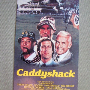 May include: A vintage movie poster for "Caddyshack" featuring actors Chevy Chase, Rodney Dangerfield, Ted Knight, Michael O'Keefe, and Bill Murray. The poster has a golf course theme with the text "The Snobs Against The Slobs!"