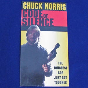May include: A movie poster for the film "Code of Silence" starring Chuck Norris. The poster features a yellow background with a black border and a red title. The poster features a photo of Chuck Norris holding a gun. The text on the poster reads "The toughest cop just got tougher."