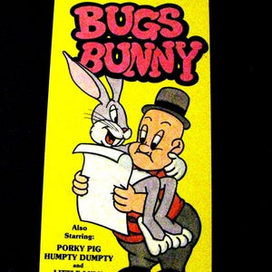 May include: A rectangular yellow VHS cover featuring Bugs Bunny and a character wearing a black hat. The text reads BUGS BUNNY, Also Starring: PORKY PIG, HUMPTY DUMPTY and LITTLE LULU. The illustration shows Bugs Bunny holding a white paper.