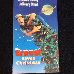 Puede incluir: Una carátula de casete de vídeo VHS con un hombre sosteniendo un árbol de Navidad. La carátula es azul con nieve blanca y el título "Ernest salva la Navidad" en letras rojas y verdes. El texto "¡Una comedia navideña como ninguna otra!" está en la parte superior de la carátula.