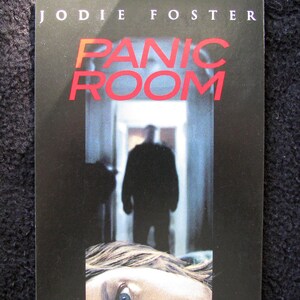 May include: A movie poster for the film "Panic Room" starring Jodie Foster. The poster features a close-up of Foster's face, looking scared, and a shadowy figure in the background.