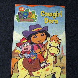 May include: A book cover featuring Dora the Explorer dressed as a cowgirl riding a horse with Boots the monkey. The book is titled "Cowgirl Dora" and says "2 Wild West Adventures!"