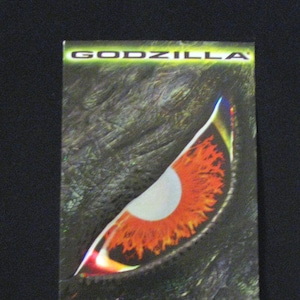 Può includere: Un primo piano di un occhio di Godzilla con un'iride rossa e arancione. L'occhio è circondato da uno sfondo scuro e testurizzato. Il testo "GODZILLA" è nella parte superiore dell'immagine e il testo "SIZE DOES MATTER" è nella parte inferiore.