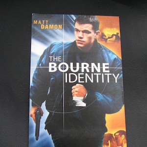 Könnte beinhalten: Ein Filmplakat für den Film "Die Bourne Identität" mit Matt Damon. Das Plakat zeigt eine Nahaufnahme von Damon als Jason Bourne, der eine Waffe hält und auf die Kamera zugeht. Der Text "Die Bourne Identität" steht in großen, fetten Buchstaben über seinem Kopf. Der Slogan "Ein Adrenalinrausch!" befindet sich am unteren Rand des Plakats.