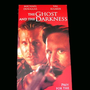 Puede incluir: Una carátula de VHS con Michael Douglas y Val Kilmer. La carátula es roja con el título "The Ghost and the Darkness" y el lema "Prey for the Hunters".