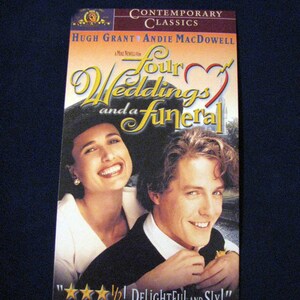 Op de afbeelding: Een VHS-cassettehoes voor de film "Vier bruiloften en een begrafenis" met Hugh Grant en Andie MacDowell. De hoes toont een close-up van de acteurs, met een rood hart en de titel van de film in een gestileerd lettertype.