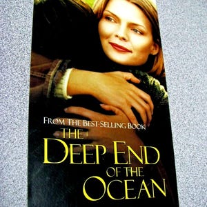 May include: A paperback book cover for The Deep End of the Ocean featuring Michelle Pfeiffer. The cover shows a person with blonde hair embracing another person. The title is printed in yellow text, and the subtitle reads: The search for her son was over. The search for her family was just beginning.