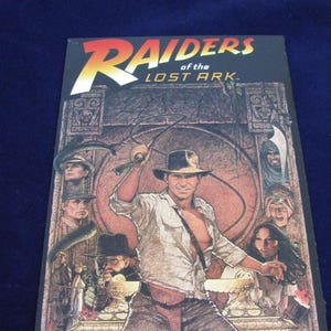 May include: A vintage movie poster for Raiders of the Lost Ark. The poster features a scene from the movie with Indiana Jones holding a whip and surrounded by other characters. The poster is black with a brown image and the title "Raiders of the Lost Ark" in yellow and red letters.